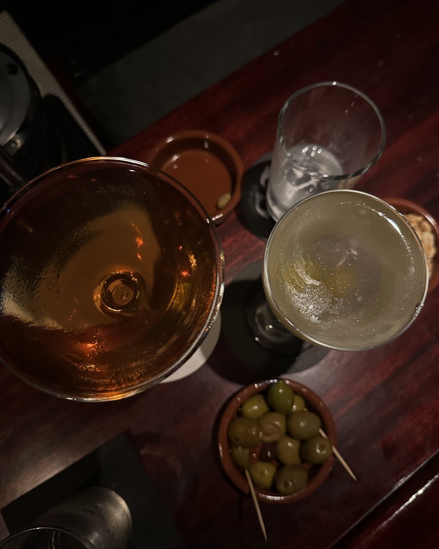 Two cocktails and a bowl of olives on a dark wooden bar, seen from above